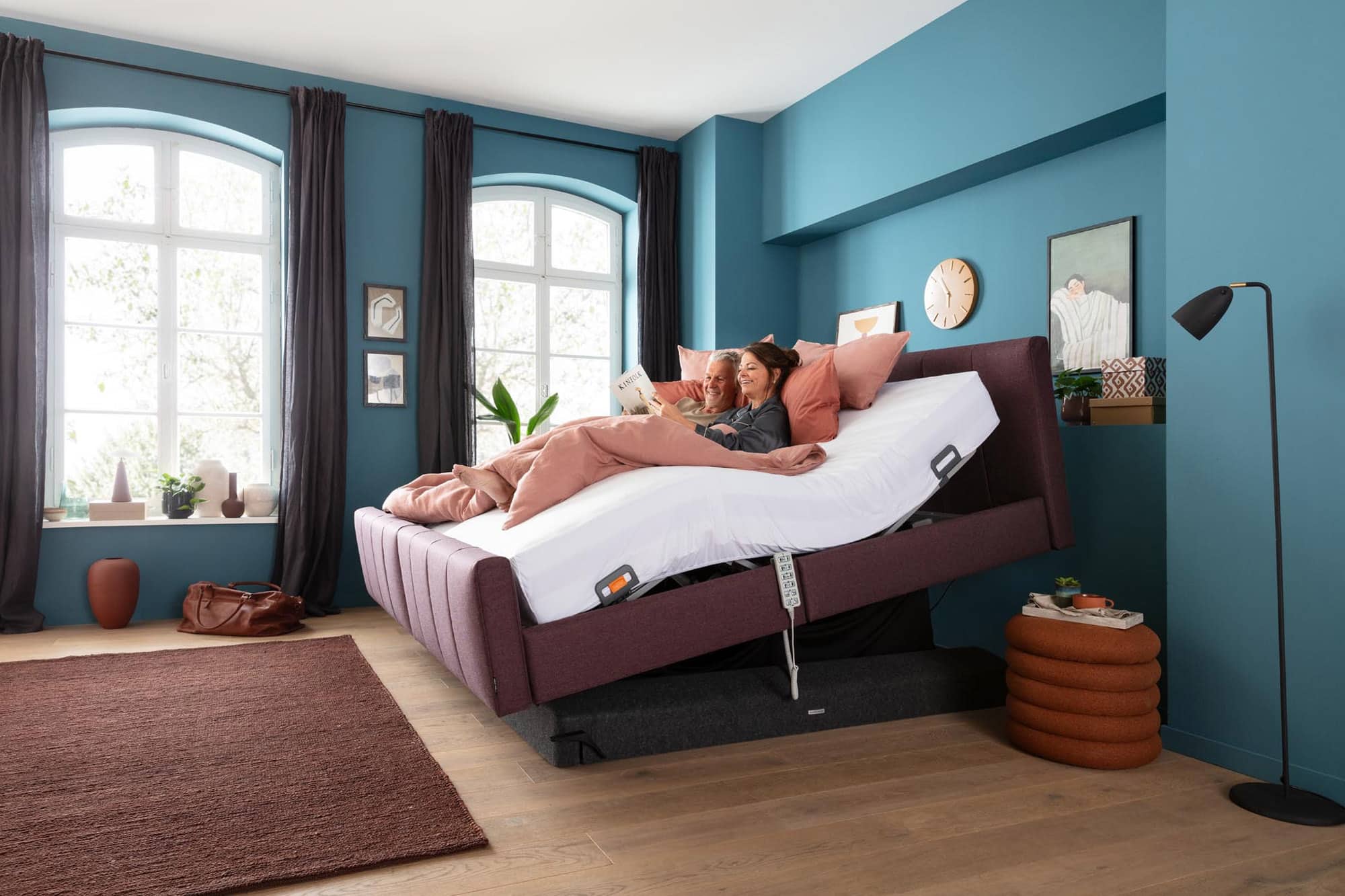 Two people relax in a modern bedroom on an adjustable bed with raised head and foot sections, reading and smiling. The room has blue walls, large windows, plants, and cozy decor.