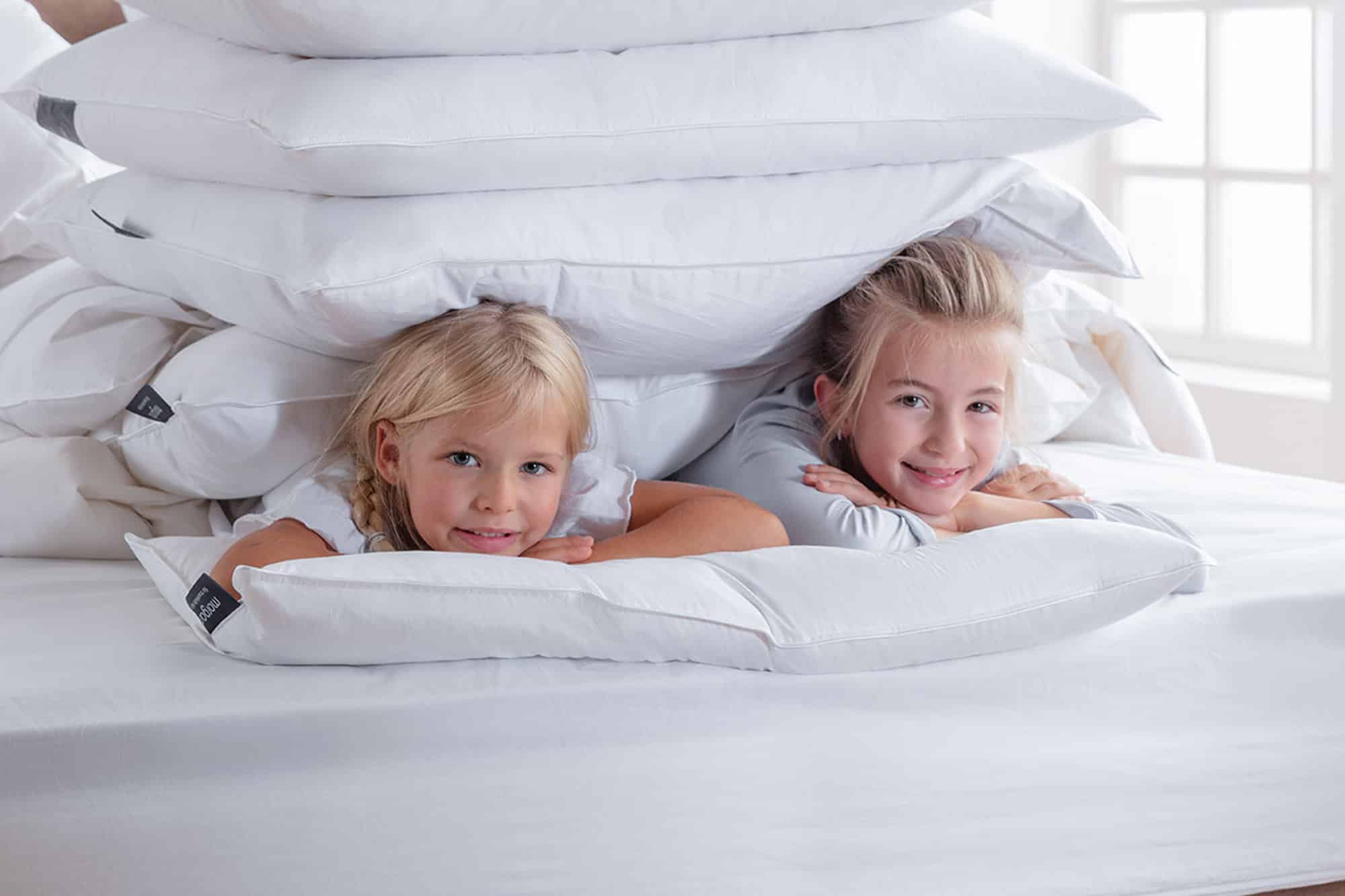 Two smiling children lie on a bed beneath a stack of white pillows, with their arms resting on top of a pillow and sunlight streaming through a window in the background.